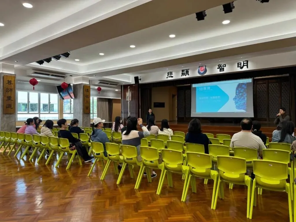 學校禮堂內，家長坐在觀眾席聆聽講座，大螢幕投影「AI 教育」主題簡報與數位人像圖像，講者持麥克風於台前分享。
