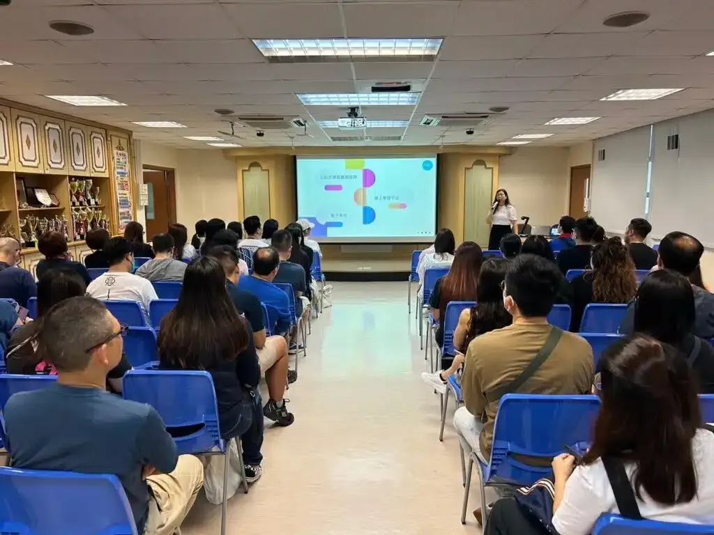 課室或小型禮堂內舉行家長講座，家長坐於藍色座椅面向講台，女講者持麥於投影幕前講解。